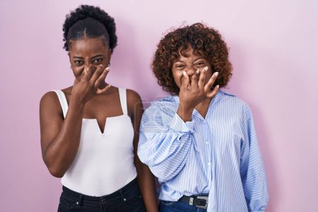 Photo for Two african women standing over pink background smelling something stinky and disgusting, intolerable smell, holding breath with fingers on nose. bad smell - Royalty Free Image