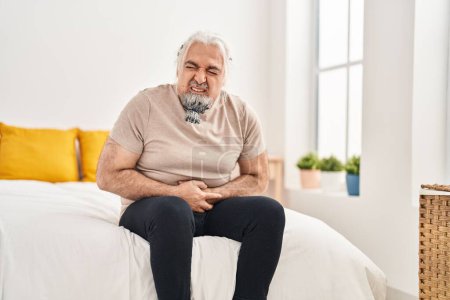 Téléchargez les photos : Moyen âge homme aux cheveux gris souffrant de maux de ventre assis sur le lit dans la chambre - en image libre de droit