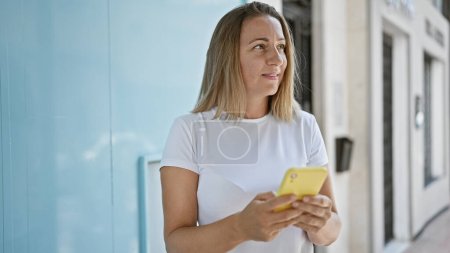 Photo for Beautiful blonde adult woman happily using her smartphone outdoors, touching screen, typing a cheerful text message on city street. - Royalty Free Image