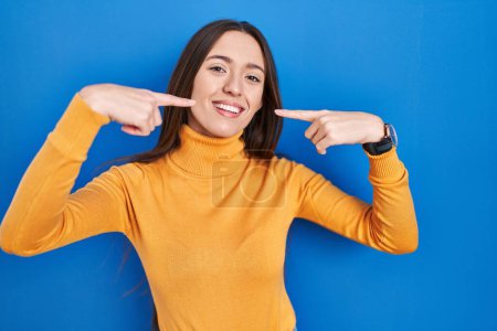 Téléchargez les photos : Jeune femme brune debout sur fond bleu souriant gai montrant et pointant avec les doigts dents et la bouche. concept de santé dentaire. - en image libre de droit