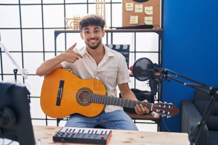 Foto de Hombre árabe con barba tocando la guitarra clásica en el estudio de música señalando con el dedo a uno mismo sonriendo feliz y orgulloso - Imagen libre de derechos