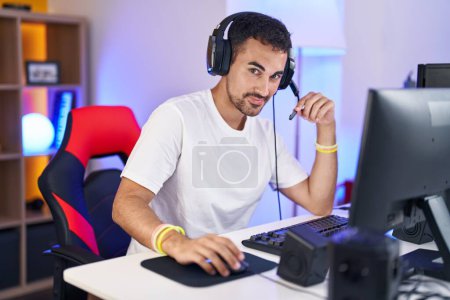 Foto de Joven streamer hispano sonriendo confiado sentado en la mesa en la sala de juegos - Imagen libre de derechos