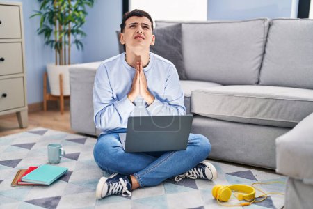 Photo for Non binary person studying using computer laptop sitting on the floor begging and praying with hands together with hope expression on face very emotional and worried. begging. - Royalty Free Image