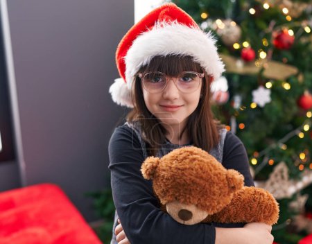 Foto de Adorable chica hispana abrazando oso de peluche de pie junto al árbol de Navidad en casa - Imagen libre de derechos