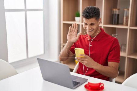 Photo for Young hispanic man using laptop and doing video call with smartphone looking positive and happy standing and smiling with a confident smile showing teeth - Royalty Free Image