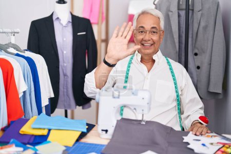 Photo for Middle age man with grey hair dressmaker using sewing machine waiving saying hello happy and smiling, friendly welcome gesture - Royalty Free Image