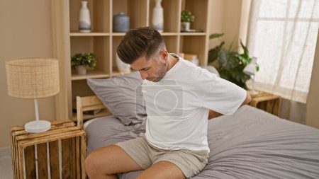 Téléchargez les photos : Inquiet, jeune homme hispanique souffrant d'une blessure au dos angoissante, allongé sur son lit de chambre, touchant malheureux son dos douloureux avec ses mains - en image libre de droit