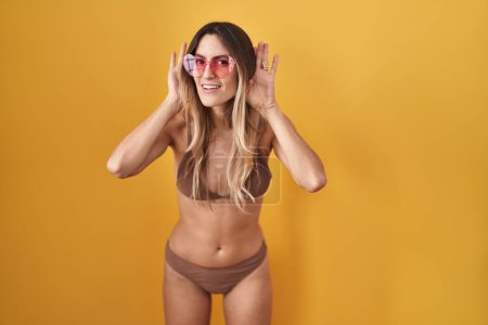Foto de Mujer hispana joven con bikini sobre fondo amarillo tratando de escuchar ambos gestos de las manos en la oreja, curiosa de chismes. problema auditivo, sordo - Imagen libre de derechos