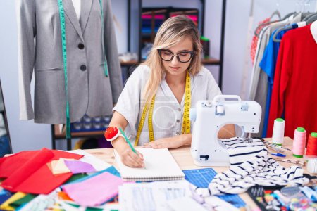 Photo for Young blonde woman tailor drawing on notebook at tailor shop - Royalty Free Image