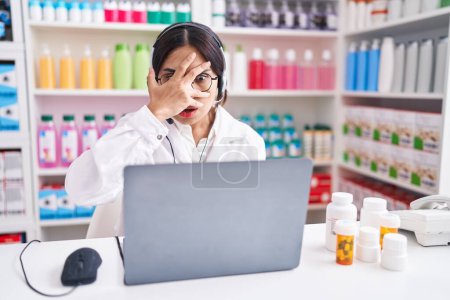 Téléchargez les photos : Jeune femme arabe travaillant à la pharmacie pharmacie en utilisant un ordinateur portable jetant un coup d'oeil dans le visage et les yeux de choc couvrant avec la main, regardant à travers les doigts avec une expression embarrassée. - en image libre de droit