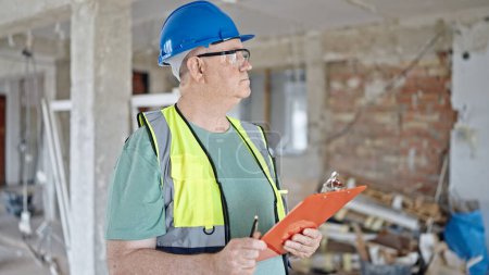 Foto de Constructor de hombre de pelo gris de mediana edad sosteniendo portapapeles mirando a su alrededor en el sitio de construcción - Imagen libre de derechos