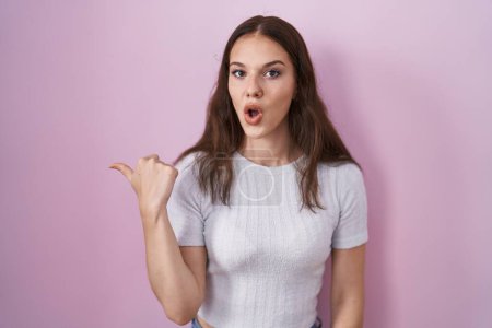 Photo for Young hispanic girl standing over pink background surprised pointing with hand finger to the side, open mouth amazed expression. - Royalty Free Image