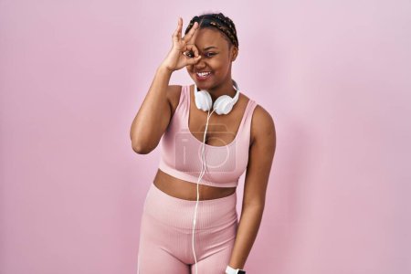 Photo for African american woman with braids wearing sportswear and headphones doing ok gesture with hand smiling, eye looking through fingers with happy face. - Royalty Free Image
