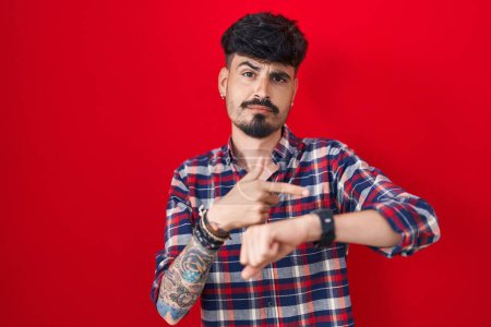 Foto de Joven hombre hispano con barba de pie sobre fondo rojo con prisa apuntando a ver el tiempo, la impaciencia, molesto y enojado por retraso plazo - Imagen libre de derechos
