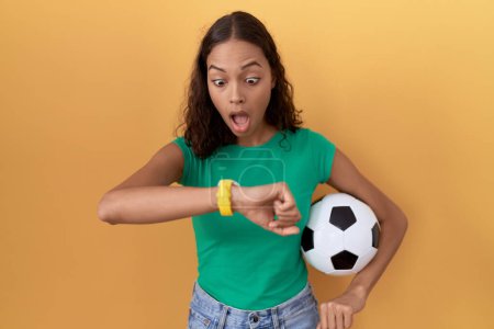 Téléchargez les photos : Jeune femme hispanique tenant le ballon en regardant le temps de la montre inquiet, peur de se faire tard - en image libre de droit