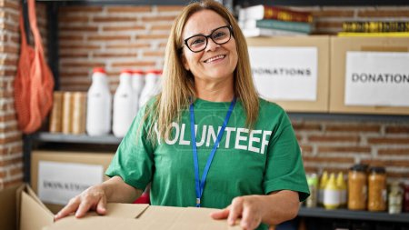Foto de Mujer hispana de mediana edad voluntaria sonriendo confiada de pie en el centro de caridad - Imagen libre de derechos