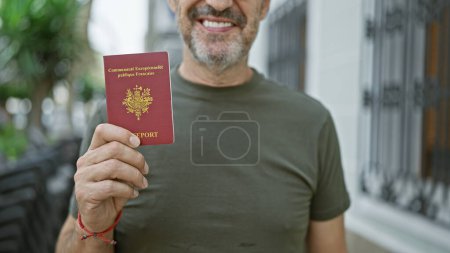 Téléchargez les photos : Homme confiant d'âge moyen aux cheveux gris affiche joyeusement son passeport français dans une rue urbaine, prêt pour son aventure passionnante de vacances en Europe - en image libre de droit