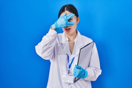 Foto de Mujer joven china que trabaja en el laboratorio científico espiando en shock cubriendo la cara y los ojos con la mano, mirando a través de los dedos con expresión avergonzada. - Imagen libre de derechos