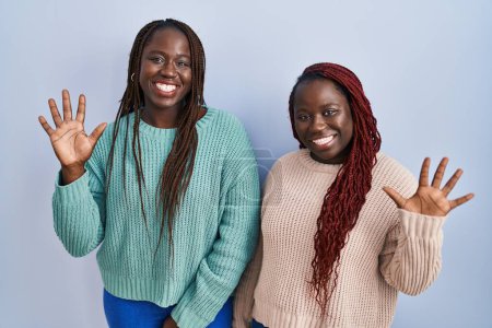 Téléchargez les photos : Deux femmes africaines debout sur fond bleu montrant et pointant vers le haut avec les doigts numéro dix tout en souriant confiant et heureux. - en image libre de droit
