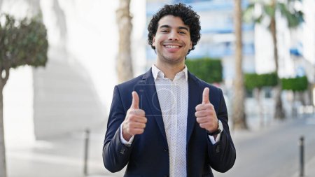 Foto de Joven trabajador de negocios latino sonriendo confiado haciendo un gesto de pulgares hacia arriba en la calle - Imagen libre de derechos