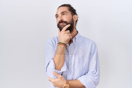Foto de Hombre hispano con barba vistiendo camisa casual mirando confiado a la cámara sonriendo con los brazos cruzados y la mano levantada en la barbilla. pensar positivamente. - Imagen libre de derechos