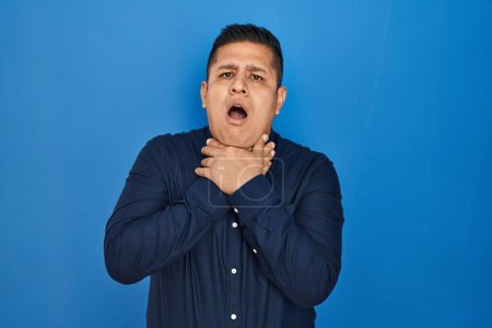 Photo for Hispanic young man standing over blue background shouting and suffocate because painful strangle. health problem. asphyxiate and suicide concept. - Royalty Free Image