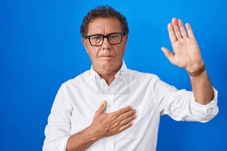 Foto de Hombre hispano de mediana edad de pie sobre fondo azul jurando con la mano en el pecho y la palma abierta, haciendo un juramento de lealtad promesa - Imagen libre de derechos