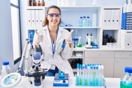 Photo for Young hispanic woman working at scientist laboratory success sign doing positive gesture with hand, thumbs up smiling and happy. cheerful expression and winner gesture. - Royalty Free Image