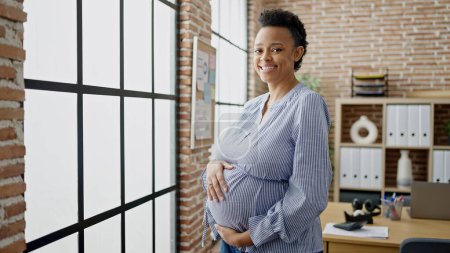 Foto de Trabajadora de negocios embarazada joven sonriendo confiada tocando el vientre en la oficina - Imagen libre de derechos