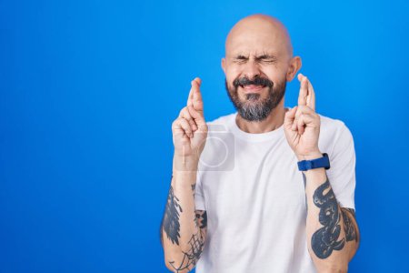 Photo for Hispanic man with tattoos standing over blue background gesturing finger crossed smiling with hope and eyes closed. luck and superstitious concept. - Royalty Free Image