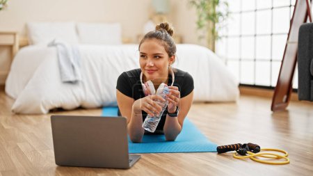 Photo for Young beautiful hispanic woman wearing sportswear lying on yoga mat holding bottle of water at bedroom - Royalty Free Image