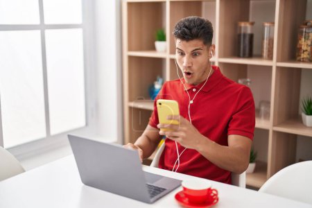 Photo for Young hispanic man using laptop and doing video call with smartphone scared and amazed with open mouth for surprise, disbelief face - Royalty Free Image