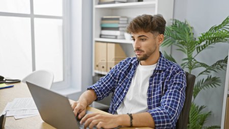 Photo for Handsome young arab man, a relaxed yet serious and elegant business pro, working hard in his indoor office space, concentrating on his laptop for success - Royalty Free Image
