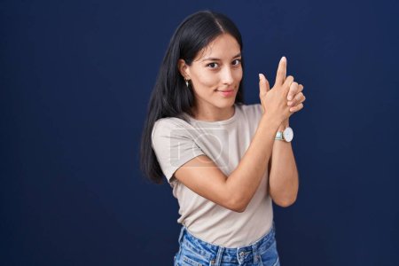 Téléchargez les photos : Jeune femme hispanique debout sur fond bleu tenant une arme symbolique avec geste de la main, jouant à tuer des armes à feu, visage en colère - en image libre de droit