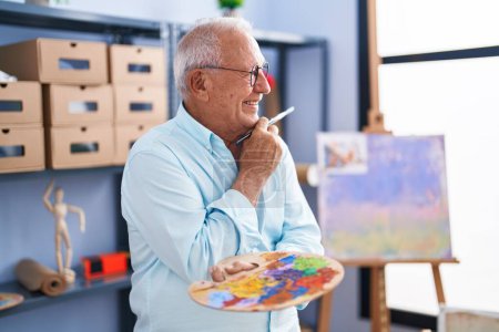 Foto de Senior grey-haired man artist smiling confident holding paintbrush and palette at art studio - Imagen libre de derechos