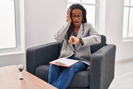Téléchargez les photos : Jeune afro-américain avec des tresses travaillant au bureau de consultation regardant le temps de la montre inquiet, peur de se retrouver en retard - en image libre de droit