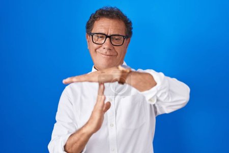 Foto de Hombre hispano de mediana edad de pie sobre fondo azul haciendo un gesto de tiempo fuera con las manos, rostro frustrado y serio - Imagen libre de derechos