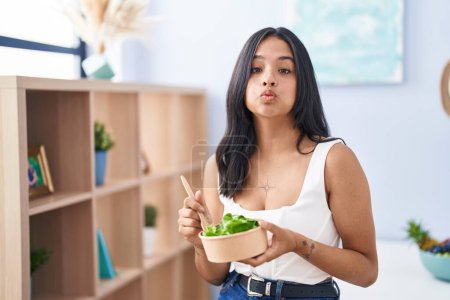 Photo for Brunette woman eating a salad at home puffing cheeks with funny face. mouth inflated with air, catching air. - Royalty Free Image