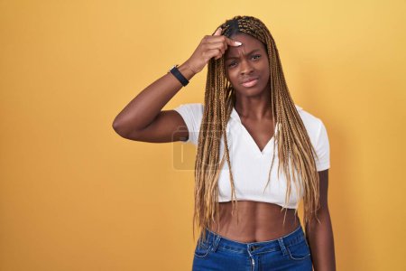 Photo for African american woman with braided hair standing over yellow background pointing unhappy to pimple on forehead, ugly infection of blackhead. acne and skin problem - Royalty Free Image