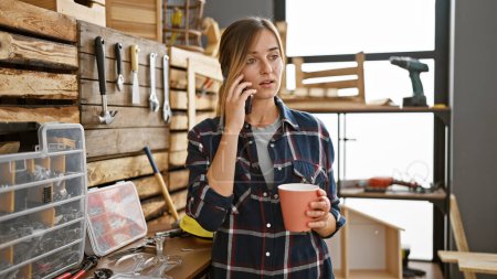 Foto de Atractiva joven mujer rubia carpintera, animada charla en el teléfono inteligente sobre el café en medio de la hermosa madera y los muebles del taller de carpintería - Imagen libre de derechos