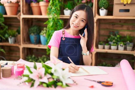 Photo for Young chinese woman florist talking on smartphone writing on notebook at flower shop - Royalty Free Image