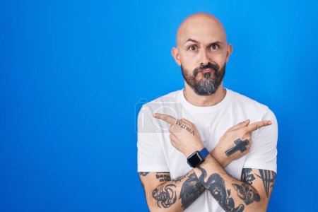 Téléchargez les photos : Homme hispanique avec des tatouages debout sur fond bleu pointant des deux côtés avec les doigts, direction différente en désaccord - en image libre de droit