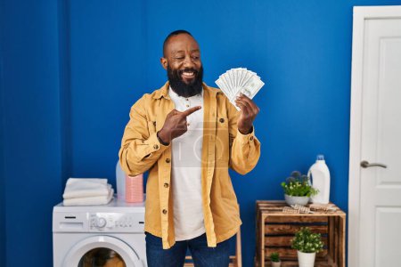 Téléchargez les photos : Africain américain homme tenant de l'argent à la buanderie souriant heureux pointant avec la main et le doigt - en image libre de droit