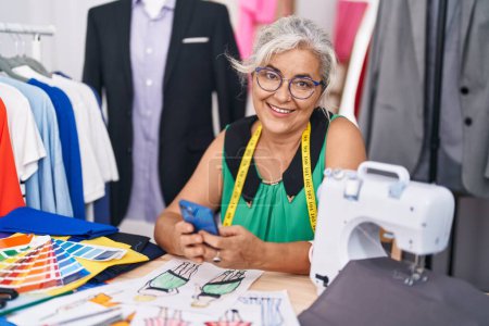 Photo for Middle age grey-haired woman tailor smiling confident using smartphone at tailor shop - Royalty Free Image