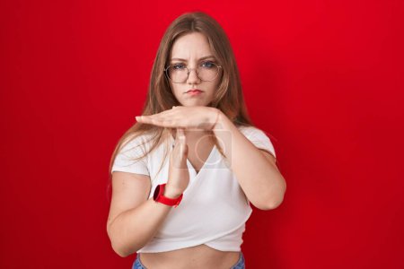 Foto de Joven mujer caucásica de pie sobre fondo rojo haciendo un gesto de tiempo fuera con las manos, rostro frustrado y grave - Imagen libre de derechos