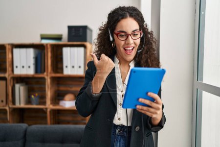 Photo for Young hispanic woman using touchpad wearing headphones pointing thumb up to the side smiling happy with open mouth - Royalty Free Image