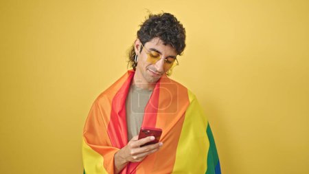 Téléchargez les photos : Jeune homme hispanique portant le drapeau arc-en-ciel à l'aide d'un smartphone sur fond jaune isolé - en image libre de droit