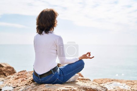 Téléchargez les photos : Femme d'âge moyen faisant de l'exercice de yoga assis sur le rocher au bord de la mer - en image libre de droit