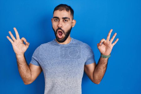 Téléchargez les photos : Moyen-Orient homme avec barbe debout sur fond bleu regardant surpris et choqué faire ok symbole d'approbation avec les doigts. expression folle - en image libre de droit