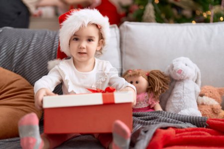 Téléchargez les photos : Adorable fille caucasienne tenant cadeau assis sur le canapé par arbre de Noël à la maison - en image libre de droit